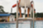 Lifeguard seated on a high chair over a pool, holding a red float. Background shows a building, trees, and calm water.