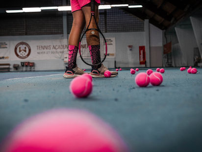 Tennis player with pink socks holds a racket on an indoor court, surrounded by scattered pink tennis balls. "Tennis Academy" sign in background.