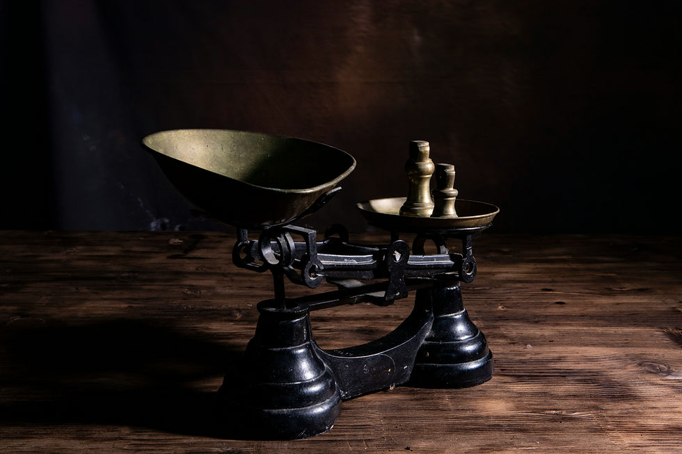 Antique brass balance scale on a wooden table, lit dramatically against a dark background, with a moody still-life feel.