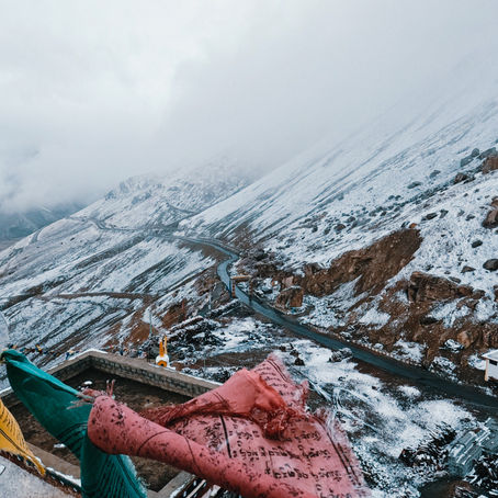 Snow-covered mountain roads winding through Ladakh, with colorful prayer flags overlooking a dramatic high-altitude Himalayan landscape.