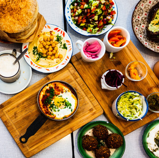 "Overhead view of a diverse breakfast board including hummus with chickpeas, falafel, pickled onions, and a mini cast-iron shakshuka."