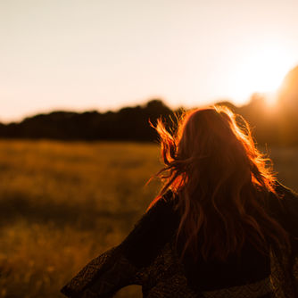 Mujer pelirroja en campo al atardecer, con cabello ondeando. Fondo de árboles oscuros y cielo cálido, creando una atmósfera serena.
