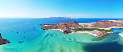 Aerial view of a coastal landscape with turquoise water, sandy beaches, rocky hills, and small boats scattered across the bay under a clear blue sky.