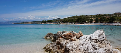 A rocky shoreline with large white rocks in the foreground, clear turquoise water, and a distant sandy beach lined with trees under a blue sky with scattered clouds.A rocky shoreline with large white rocks in the foreground, clear turquoise water, and a distant sandy beach lined with trees under a blue sky with scattered clouds.