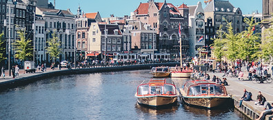 A canal in a European city with tour boats docked, people sitting along the waterfront, and historic buildings with gabled roofs in the background under a clear blue sky.