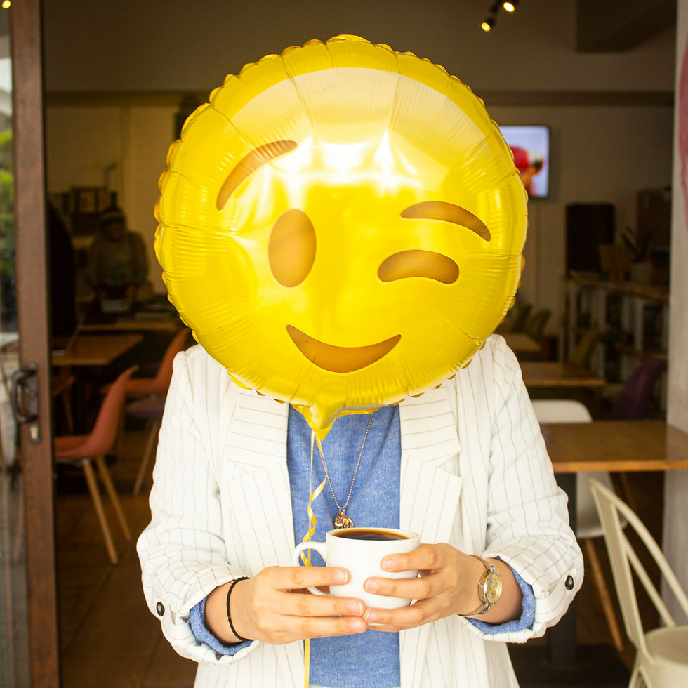 Person holds a cup while their face is obscured by a winking emoji balloon in a cafe. The setting is warm and casual with wooden tables. Represents person with high functioning depression who is masking.