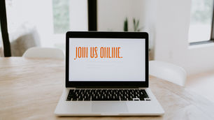 Laptop auf einem Holztisch mit der Bildschirmaufschrift „Join Us Online.“ in orangefarbener Schrift, in einem hellen, modernen Raum mit unscharfem Hintergrund.