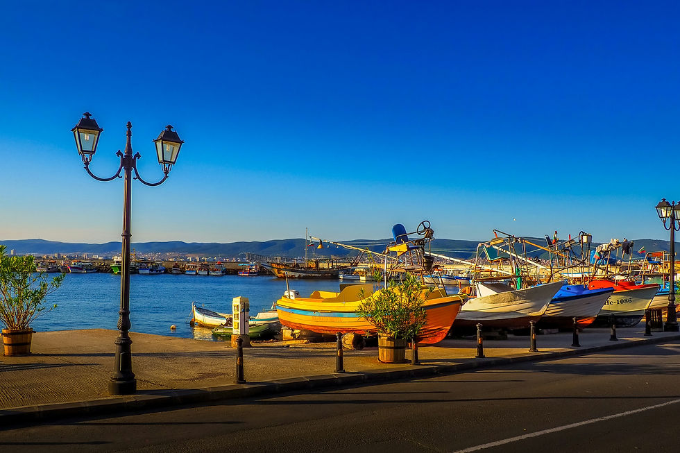 Krāsainas laivas stāv piestātnē saulainā piekrastē, fonā zilas debesis un kalni. Bulgārija