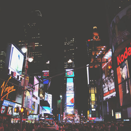 Times Square at night, bustling with people. Bright, colorful billboards display ads like Kodak and Virgin, creating a vibrant urban scene.