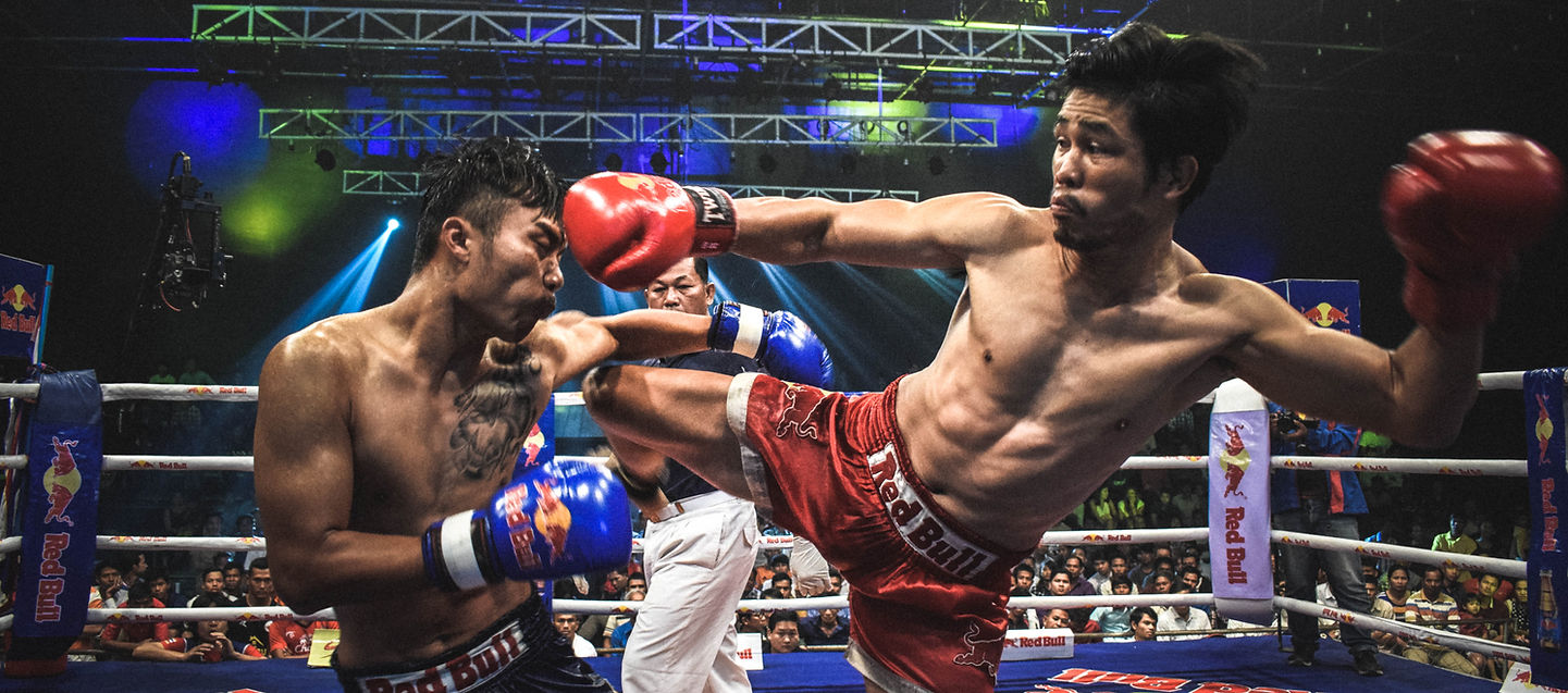 Two Muay Thai fighters in the ring mid-kick exchange under stadium lights.