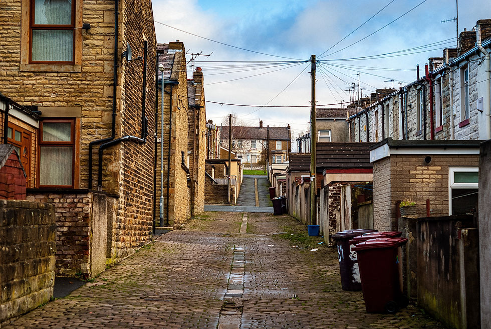 Cobblestone alley between stone houses, lined with garbage bins, leading to distant houses. Overhead wires under a cloudy sky. Quiet mood.
