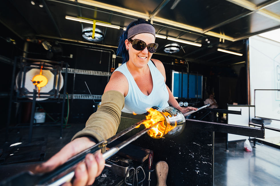 Smiling woman in sunglasses shapes molten glass with a tool in a workshop, flames visible. She's wearing a blue tank top and headband.