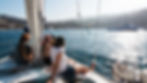 People relaxing on a sailboat in a sunny harbor, surrounded by calm blue water and moored boats, with distant mountains and a sunny sky.