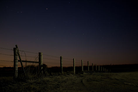 Barbed wire fence lines a dirt path under a starry night sky. The horizon glows softly, casting a tranquil, moody atmosphere.