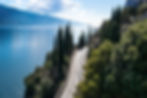 Curving road along a clear blue lake, lined with tall cypress trees and lush greenery, under a bright sky with fluffy clouds. Serene landscape.