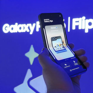 Main tenant un smartphone Samsung Galaxy Flip6 plié, avec en arrière-plan l’affichage lumineux du logo Galaxy Flip6 sur fond bleu.