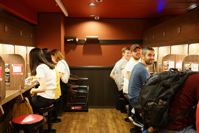 Collage gastronómico de Japón: un bowl de ramen caliente en una mesa de madera, la fachada iluminada de restaurantes callejeros, una fila de clientes comiendo en cabinas individuales estilo Ichiran, y faroles japoneses tradicionales colgando en una calle nocturna.