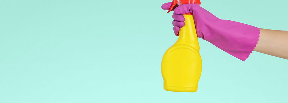 The image depicts a hand in a pink rubber glove holding a yellow spray bottle, set against a simple light teal background, symbolizing household cleaning or chores. There’s a focus on the cleaning action, suggesting an emphasis on cleanliness and maintenance.