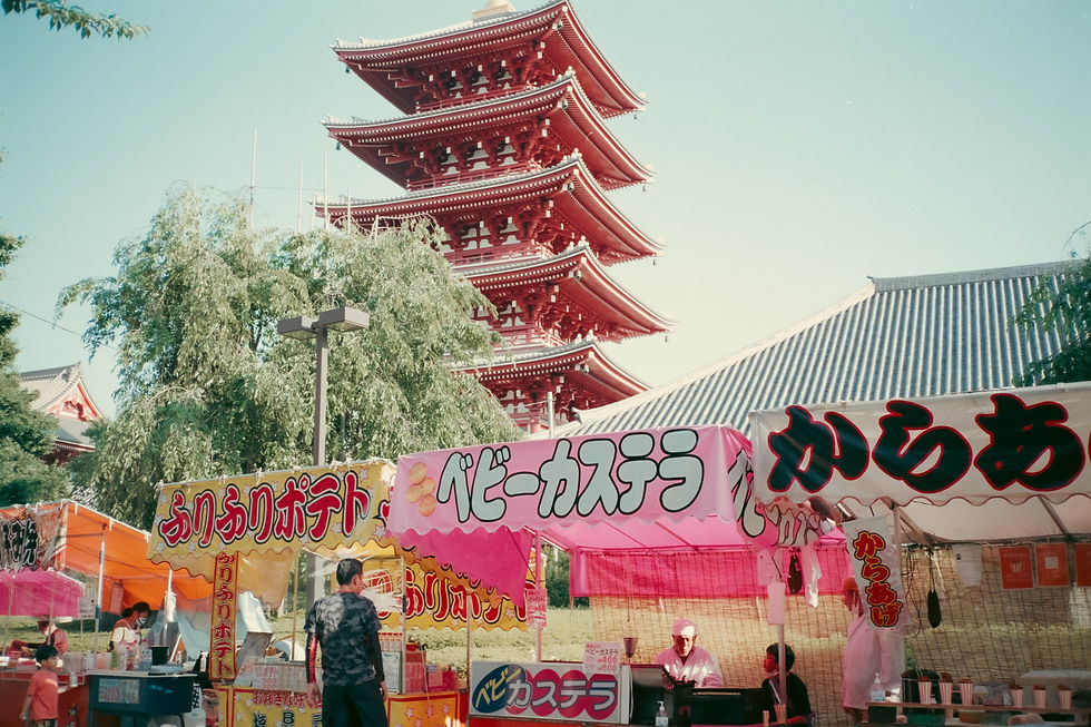 Puestos de comida con letreros coloridos en un mercado japonés. Al fondo, una pagoda roja y árboles. Ambiente animado y soleado.