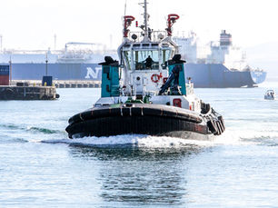 Tugboat speeding through a misty harbor, with huge cargo ships and a dock in the bright background.