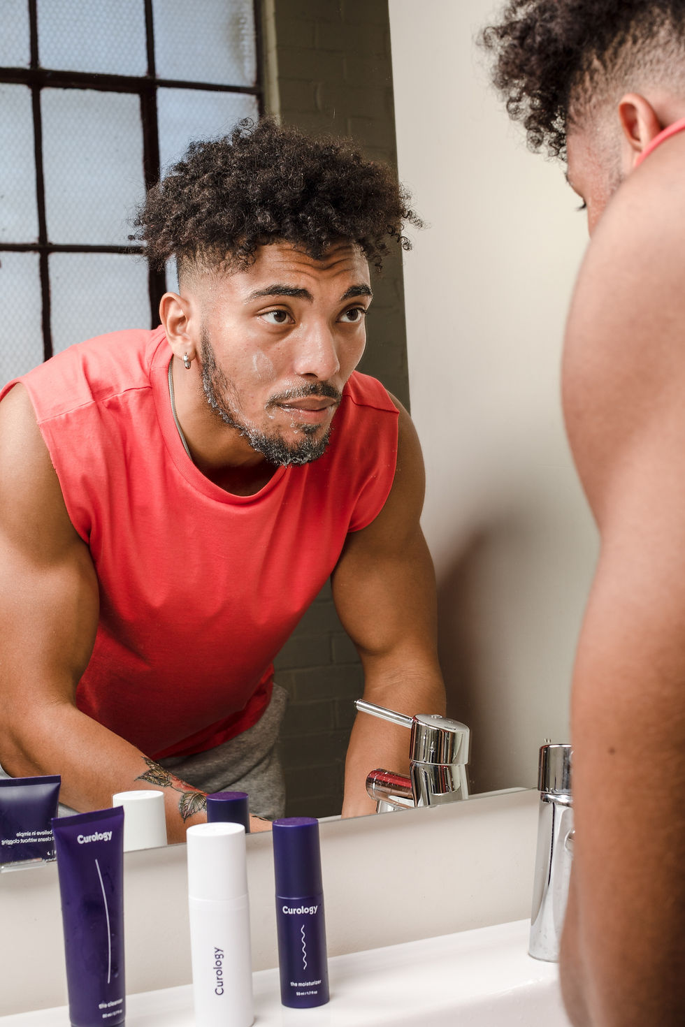 Man in a red shirt examines his face in a bathroom mirror. Skincare products are on the sink. Natural light from a window fills the room.