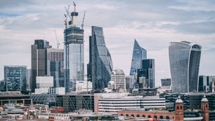 Skyline of a modern city with high-rise buildings, including uniquely shaped skyscrapers and a construction crane, under a cloudy sky.