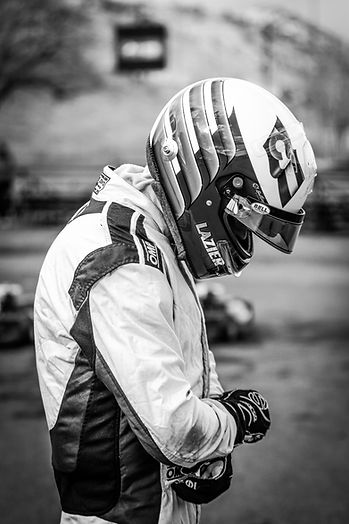 A black and white photo of a motorsport driver in all gear looking down towards their helmet