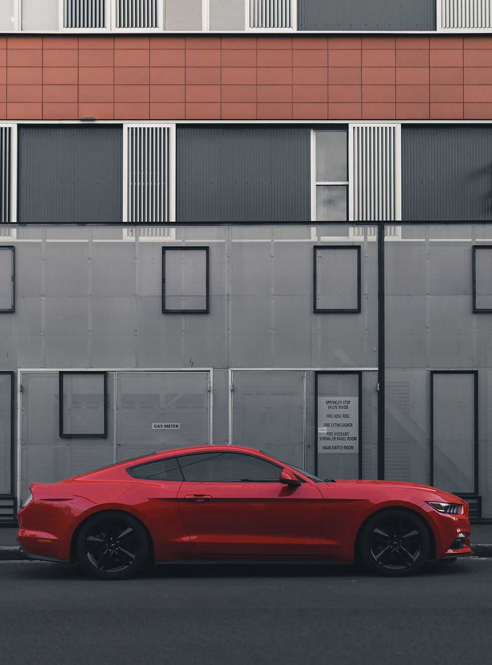 A shiny red Ford Mustang parked in front of a modern building with a gray, white, and terracotta-colored geometric facade. The car's sleek profile showcases its tinted windows against the urban backdrop.