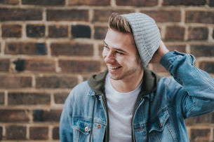 A smiling person in a gray beanie and denim jacket stands against a brick wall, hand on head, exuding a relaxed and cheerful mood.