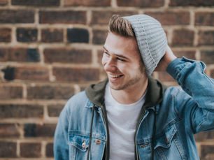 A smiling person in a gray beanie and denim jacket stands against a brick wall, hand on head, exuding a relaxed and cheerful mood.