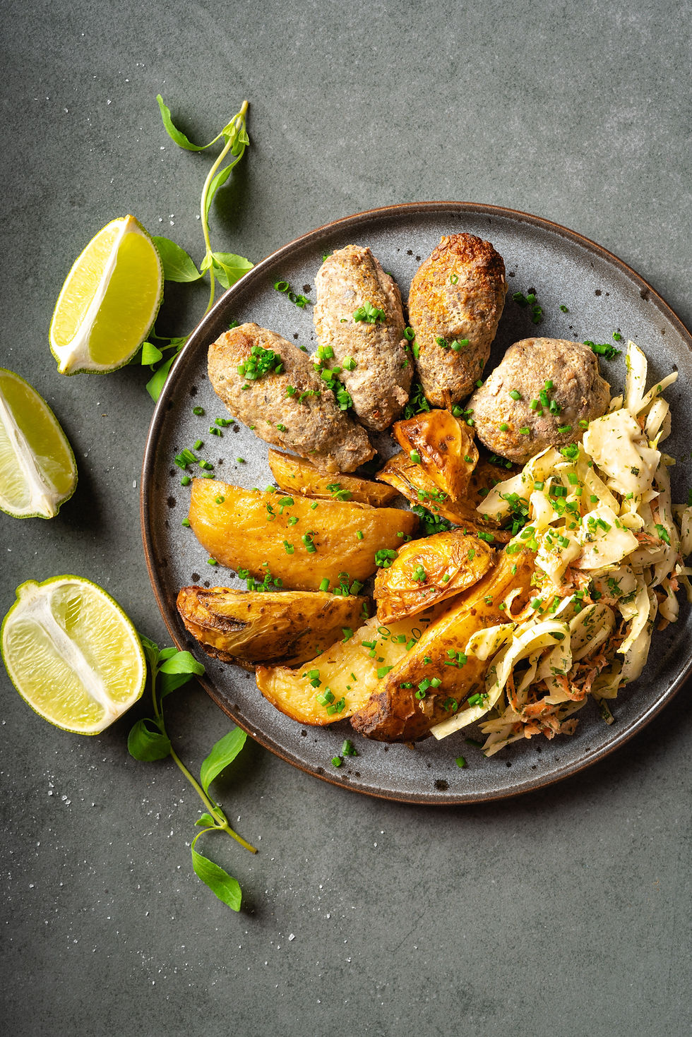 Plate with sausages, roasted potato wedges, and cabbage salad, garnished with chives. Lime wedges and fresh greens on side.