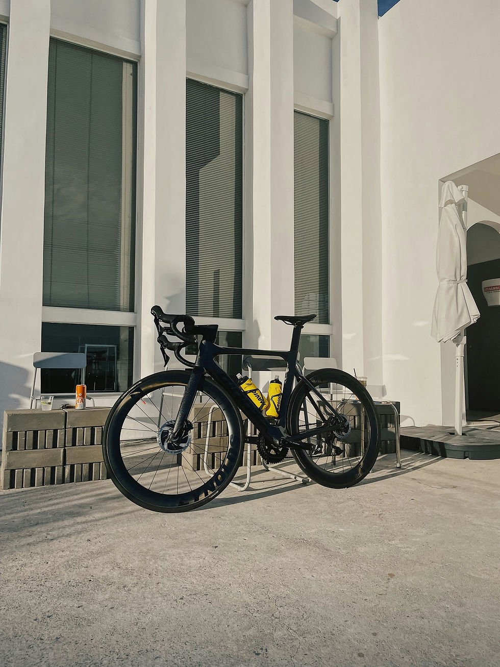A sleek black road bike with yellow water bottles is parked in front of a modern, minimalist white building, basking in the warm sunlight.