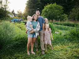 A family of five stands in a lush garden at sunset, smiling. A blue tractor is visible in the background amidst tall green plants.