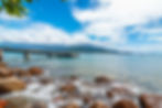 Rocky shoreline in Ilhabela with waves, wooden pier extending into turquoise sea, mountains under blue sky with clouds. Peaceful, natural setting.