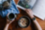 Hand with cup of tea, next to bowl of muesli, other hand about to turn page in a book