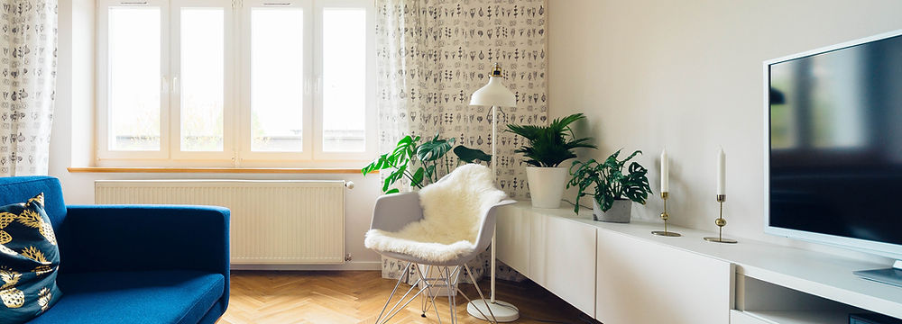 A bright, cozy living room corner featuring a white armchair with a throw blanket, surrounded by green houseplants. A blue sofa is partially visible, and the room has a herringbone wood floor, white walls, and large windows with geometric curtains.