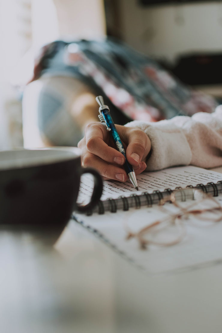 Close-up of a hand writing in a spiral notebook with a blue pen, beside a blurred mug in a soft, cozy indoor scene.