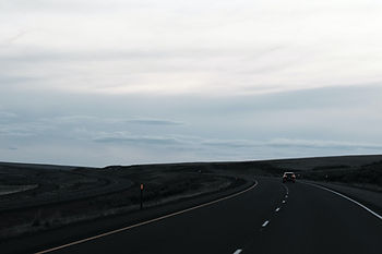 Car drives on a dark, winding highway through open plains under a cloudy gray sky, creating a quiet, moody scene.