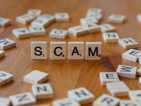 Wooden tiles spelling "SCAM" on a wooden table, surrounded by scattered letter tiles. Mood is cautionary.