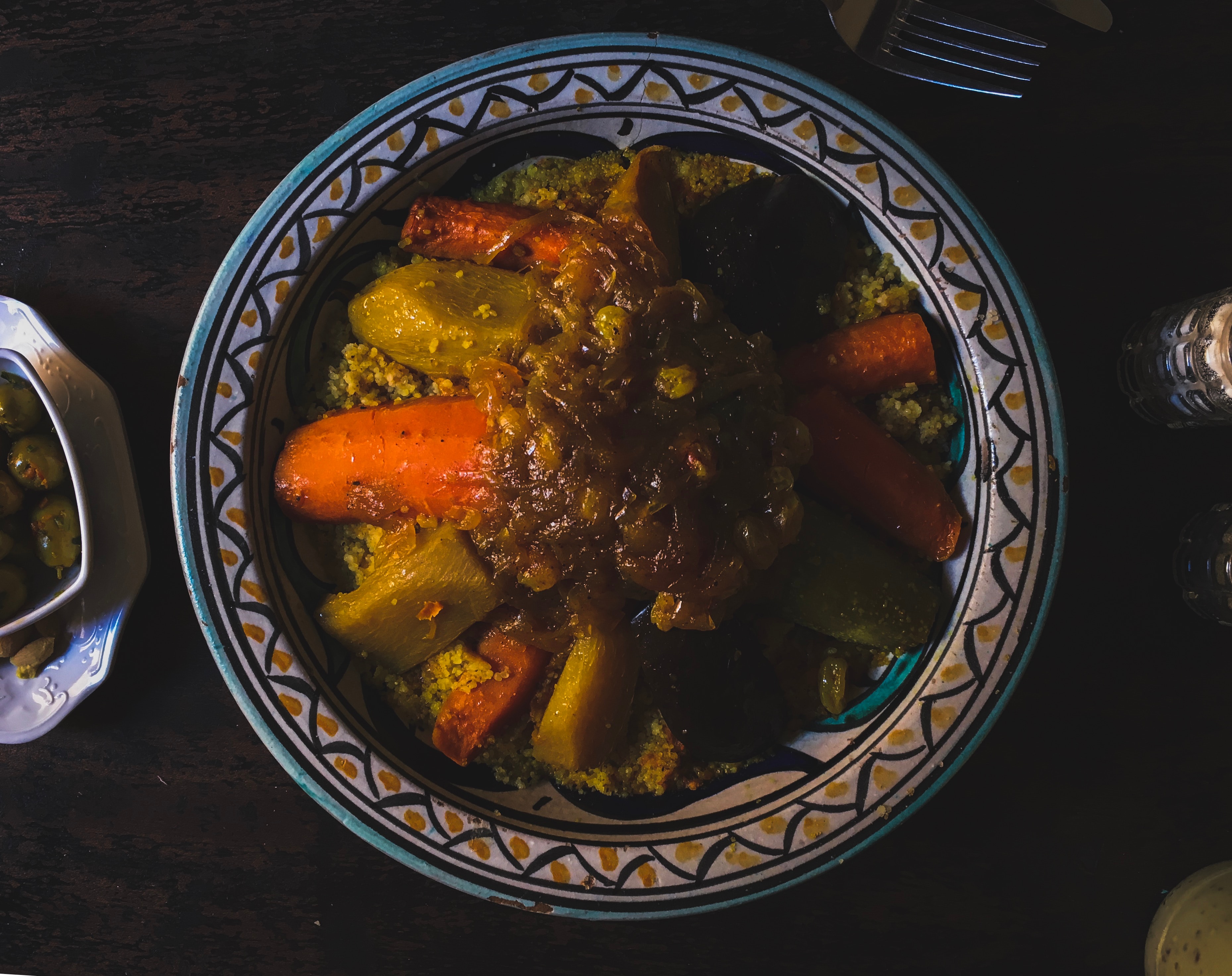 A colorful ceramic plate with couscous topped with carrots and vegetables on a dark wooden table. A small dish of olives is nearby.