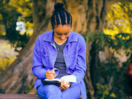 Jeune femme en veste bleue lisant et écrivant dans un cahier, assise devant un grand arbre, ambiance paisible et automnale.