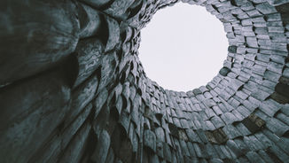 Looking up at a circular wooden structure with curved planks, revealing a pale sky. The design creates a tunnel-like effect.