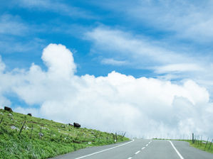 Carretera vacía entre colinas verdes bajo cielo azul con nubes blancas, paisaje tranquilo y abierto.