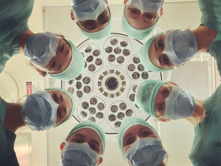A group of surgeons wearing masks and surgical caps look down toward the camera in a circle, with bright operating room lights centered above them