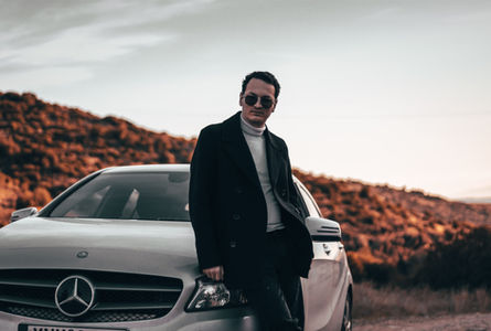 Man in sunglasses and dark coat leans against a silver Mercedes on a rocky roadside, under a pale sky with a cool, moody feel