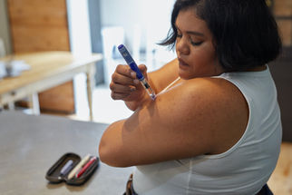 Woman in white tank top injects insulin into her arm at home. On the table, there's a case with pens. Bright room with wooden decor.