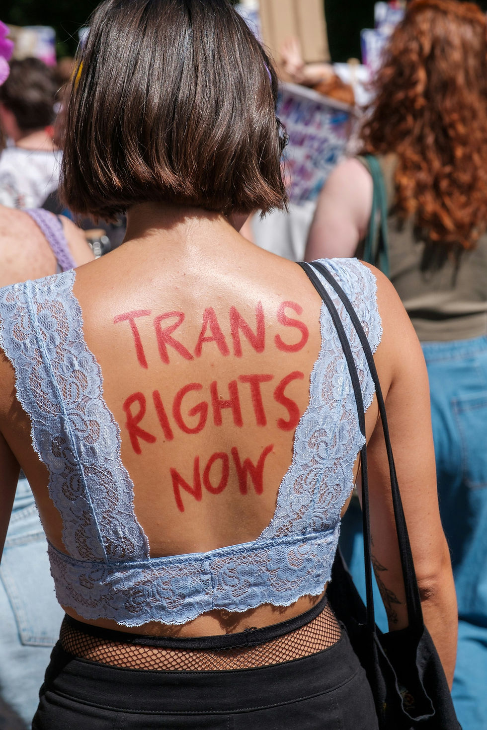 A person at a rally wears a backless lace top with the bold message "Trans Rights Now" painted in red on their back, advocating for transgender rights.