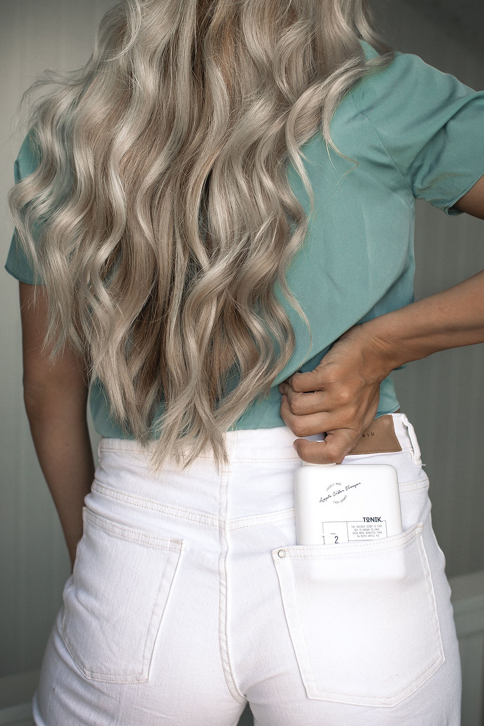 blonde wavy hair modeled from the back of a woman wearing white jeans, pulling shampoo out of the back pocket.