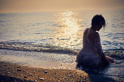 Woman in a white lace dress sits on a pebbled beach at sunset, gazing at the shimmering sea. Warm golden hues create a serene mood.