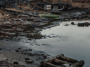 Abandoned boats by a weathered house near a rocky shore under a cloudy sky. Grass and wooden debris enhance an eerie, desolate mood.